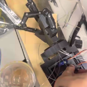 Top-down view of a robotic hand prototype mounted on a wooden base. The fingers are connected to clear strings that lead to a newly designed servo bed. A hand is adjusting one of the servo motors, which control the finger movements in response to rotations.